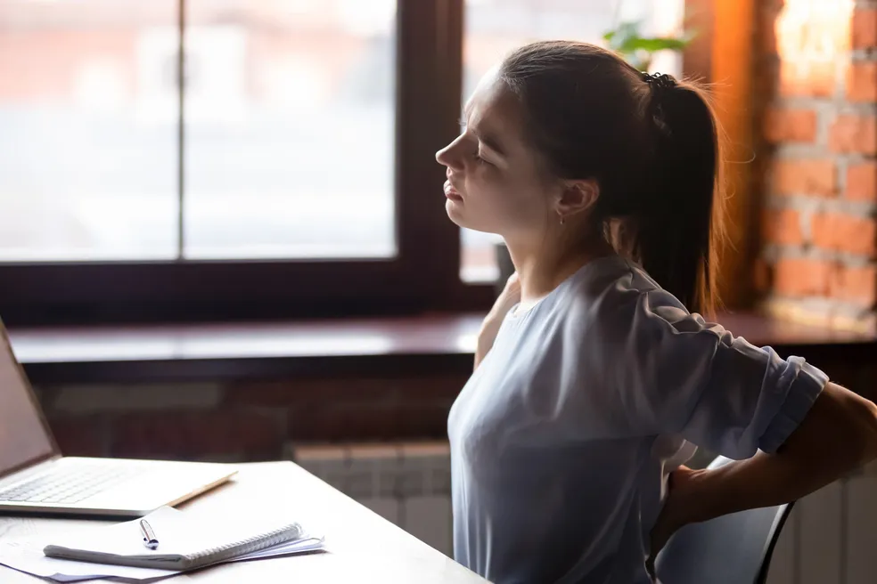 Mulher sentada à mesa de trabalho com a mão na lombar, sinalizando dor nas costas