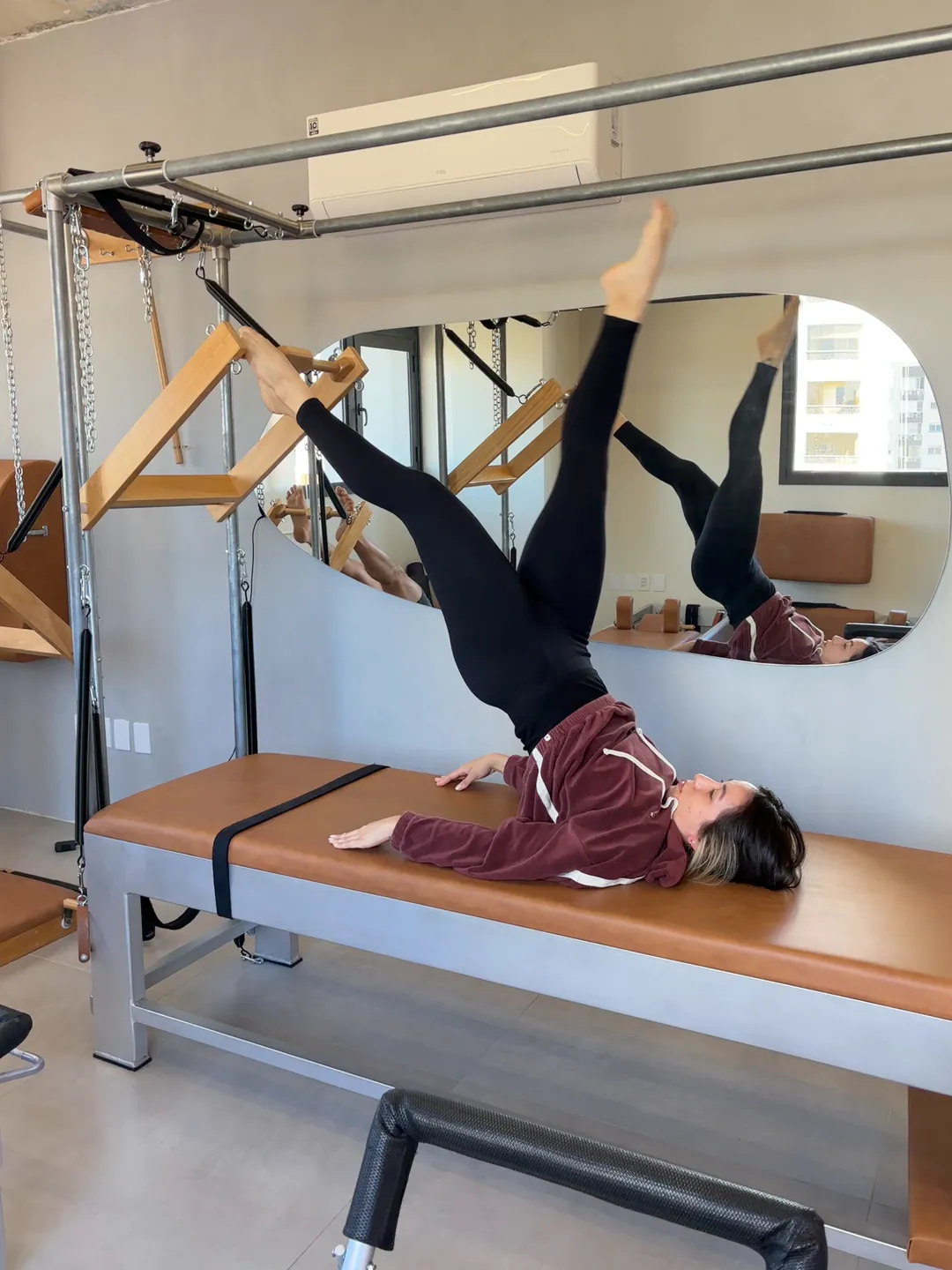 Aluna deitada na mesa do Cadillac, com um pé na barra de molas e a outra perna estendida para o alto, em aula no Hera Pilates Clássico, no Menino Deus, Porto Alegre