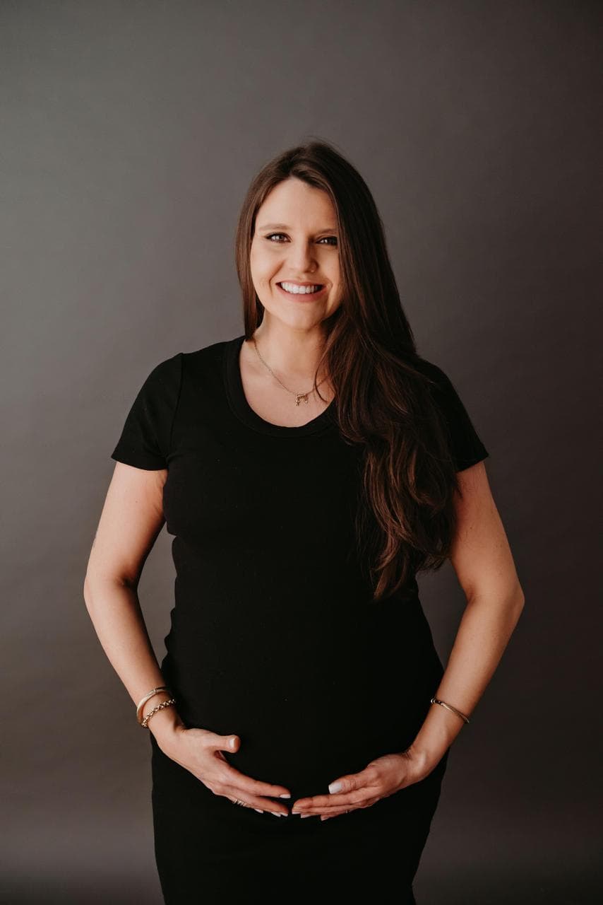 Michele Flach, fisioterapeuta do Hera Pilates Clássico em Porto Alegre, grávida, sorrindo com as mãos na barriga