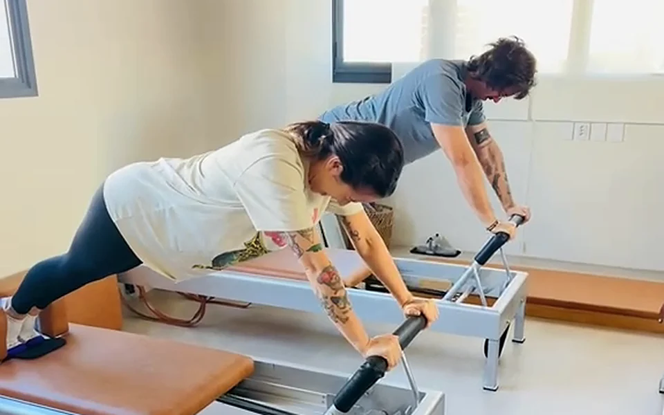 Aluna e aluno em prancha no Reformer, com as mãos na barra e os pés apoiados no carrinho, em aula de Pilates no estúdio Hera, no Menino Deus, em Porto Alegre
