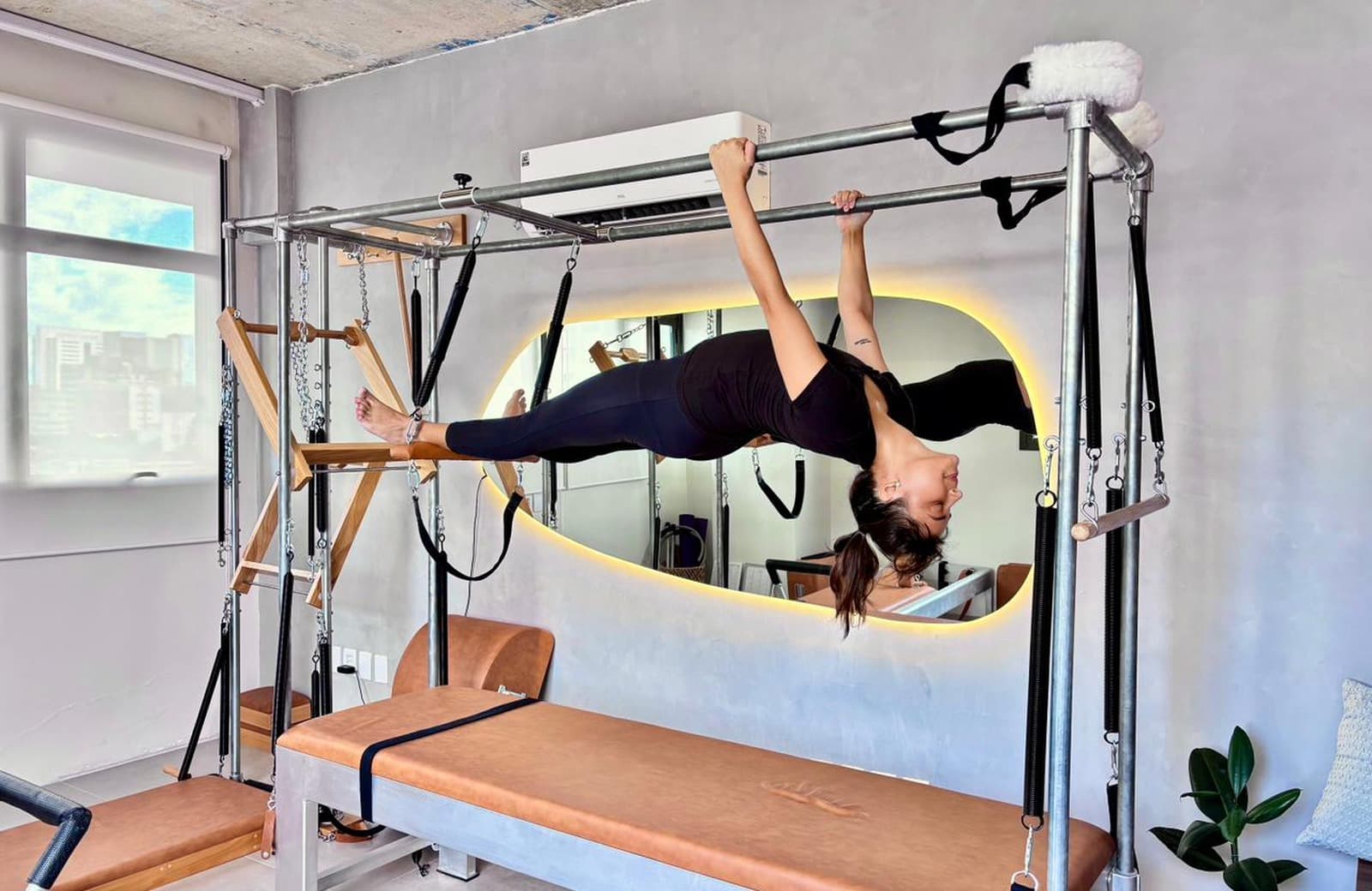 Aluna suspensa na barra do Cadillac diante do espelho de luz quente do Hera Pilates Clássico, no Menino Deus, Porto Alegre